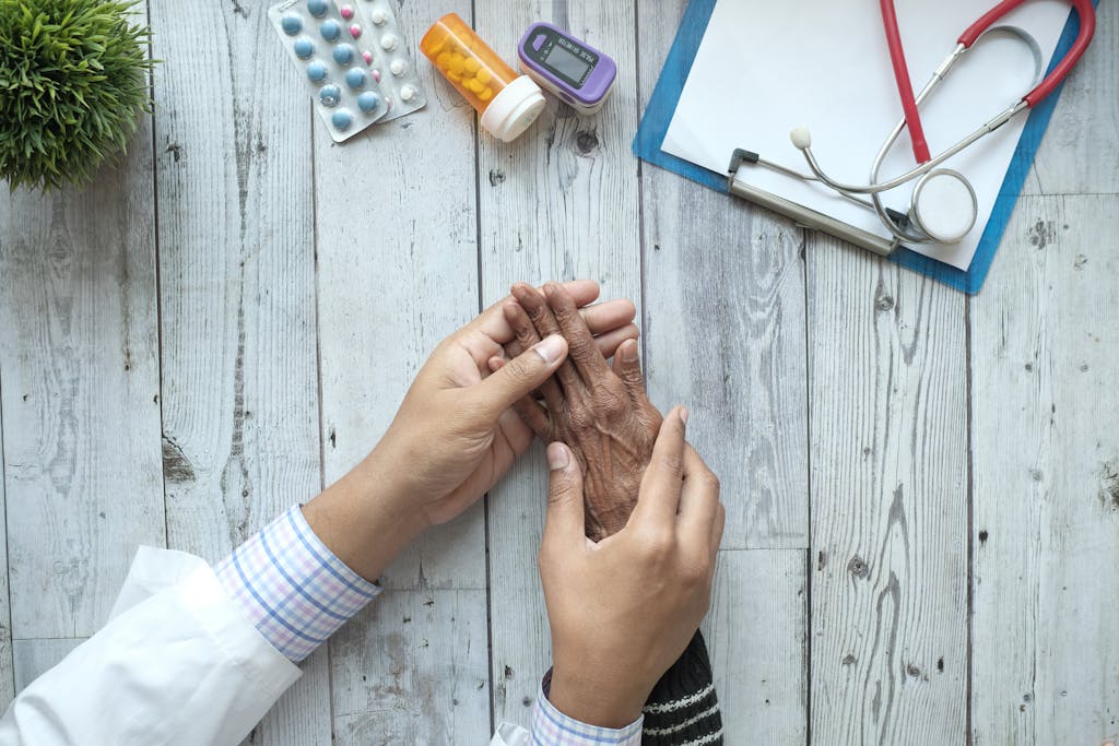 A doctor holding an elderly patient's hands on a desk with medication and stethoscope.